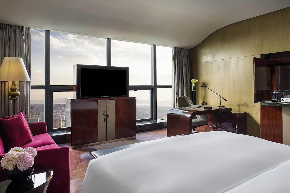 A hotel room with a large bed, a TV on a wooden cabinet, a desk with a chair, and a view of the city through large windows.