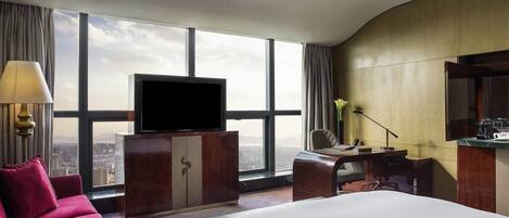 Habitación de hotel con una cama grande, un televisor sobre un mueble de madera, un escritorio con una silla y vistas a la ciudad a través de amplias ventanas.
