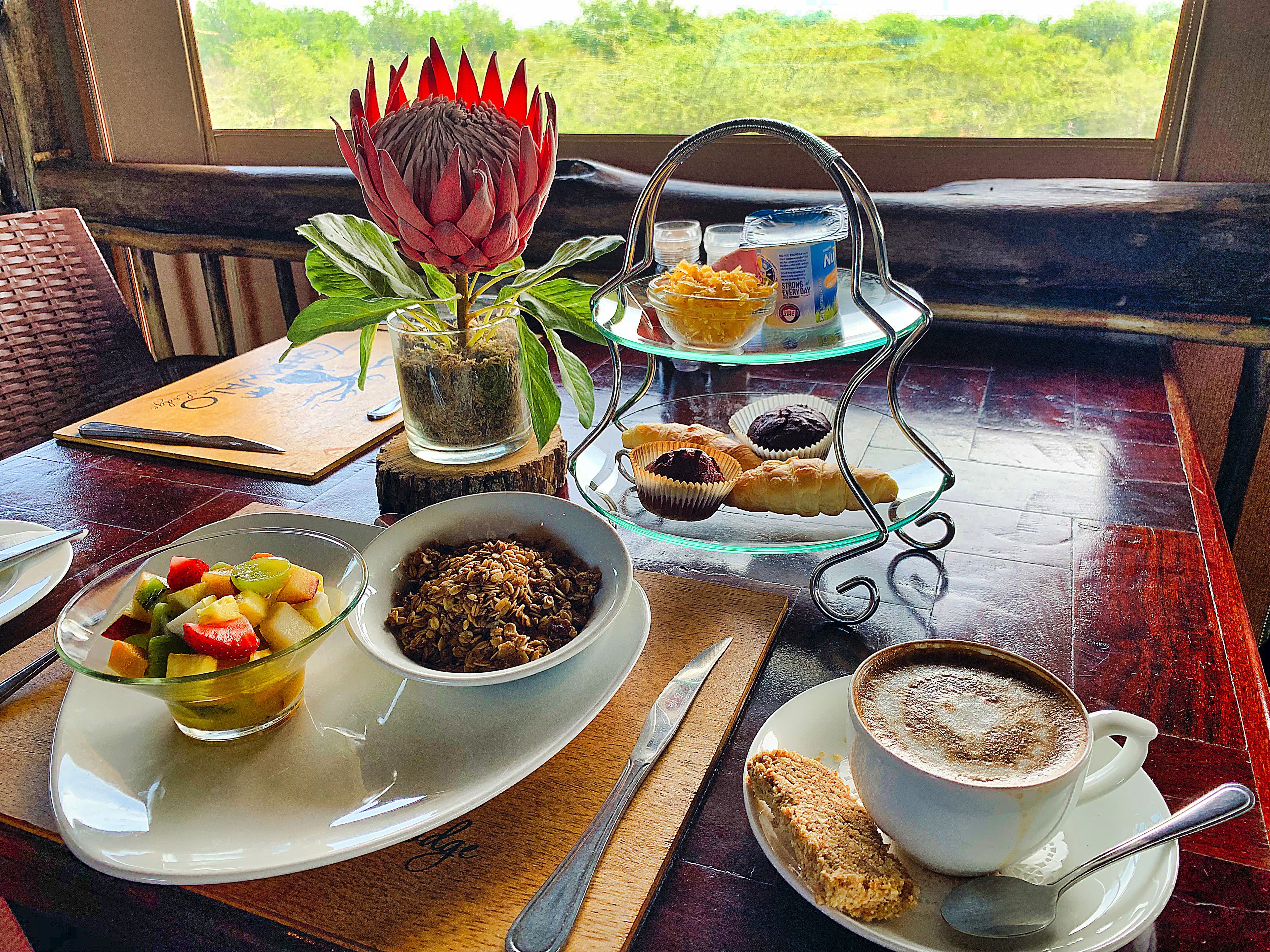 Ein Tisch reichlich gedeckt mit Obstschale, Granola, einer Kaffeetasse, Gebäck und einer Zierpflanze.