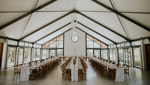 Un salón amplio con techos altos y una lámpara central, amoblado con mesas y sillas, y ventanas grandes a los lados.
