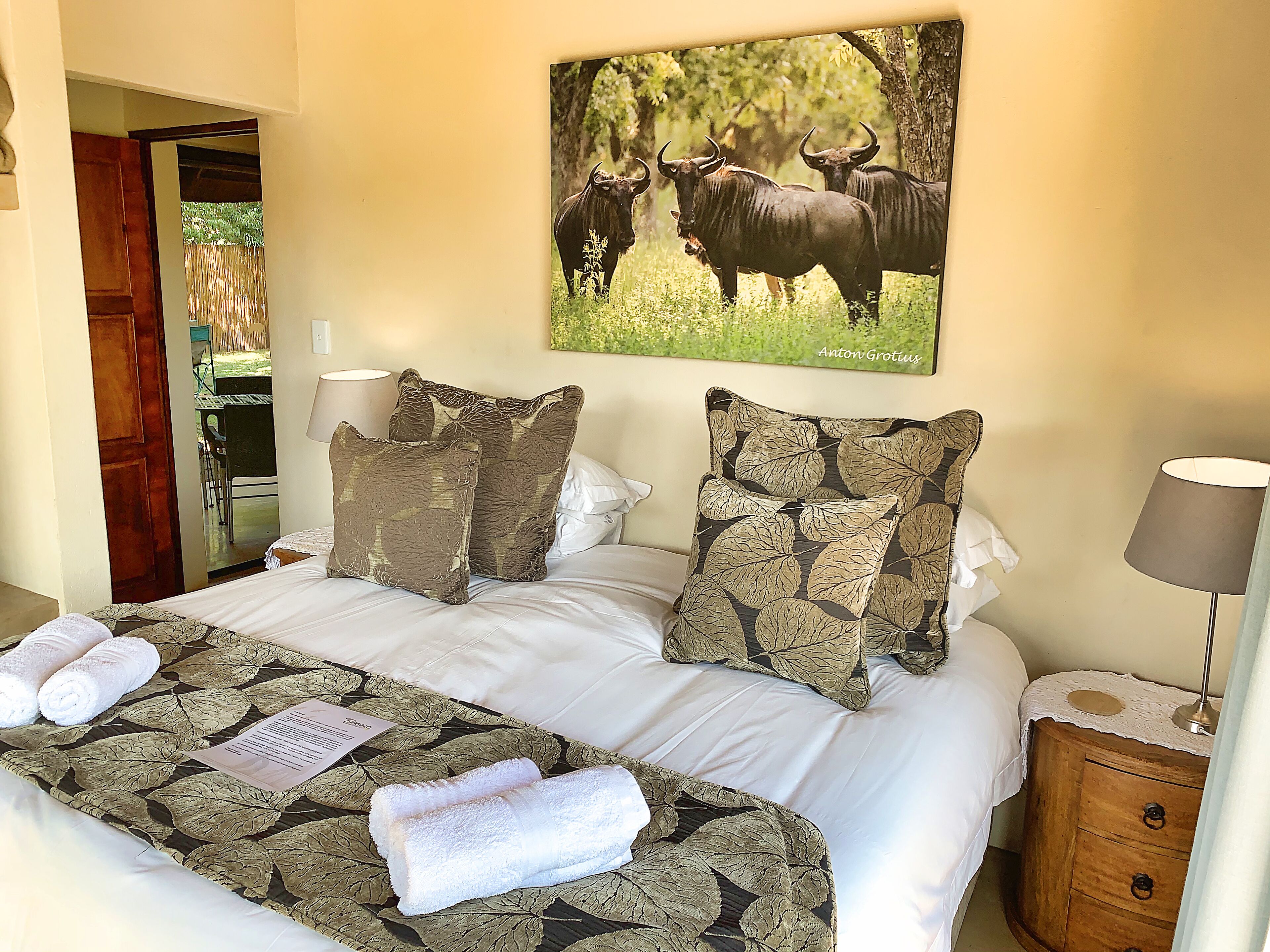 A hotel room with a bed, pillows, a nightstand, a lamp, and a painting of wildebeests.