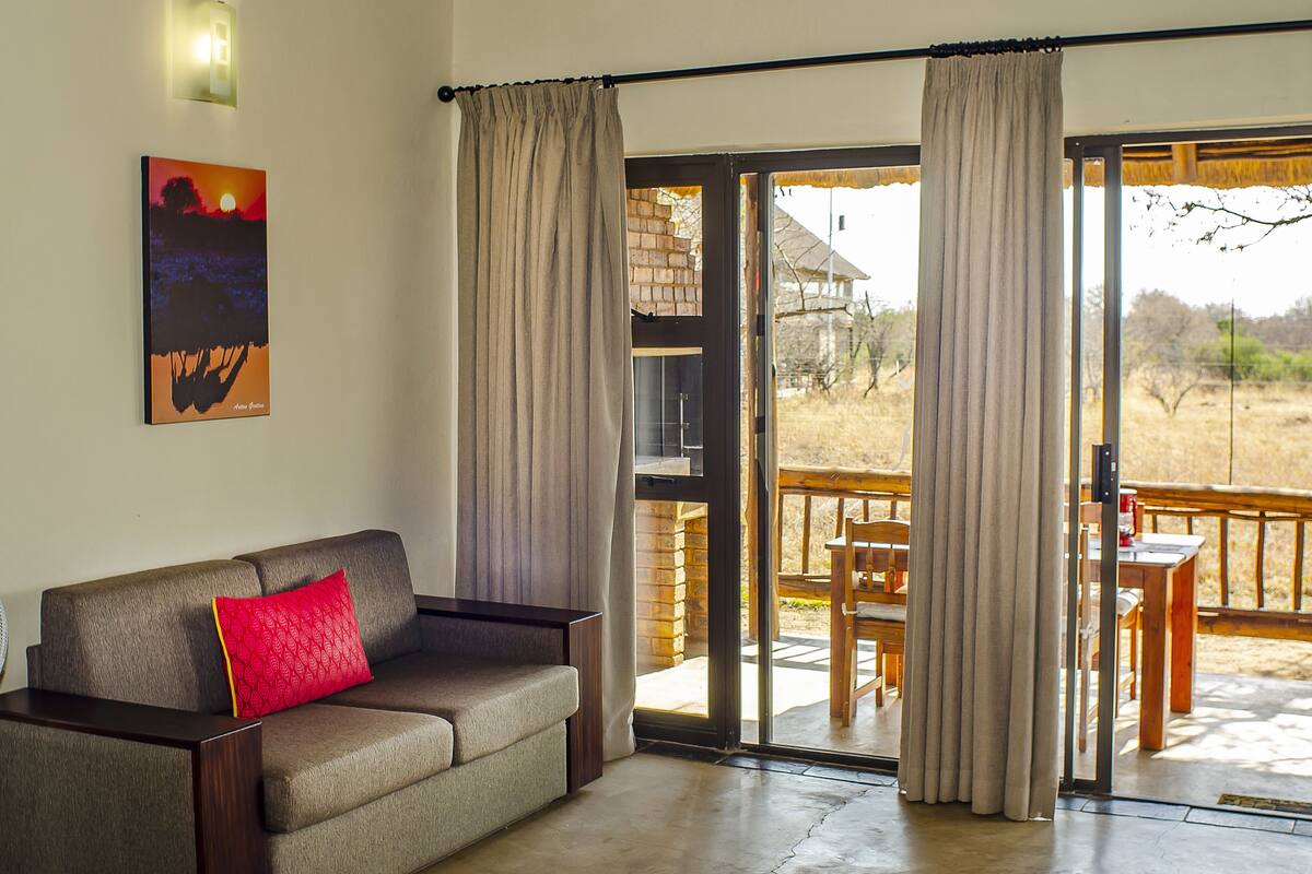 Habitación con sofá, mesa de comedor y puerta corrediza de vidrio que da a un espacio exterior con terraza de madera y sillas.