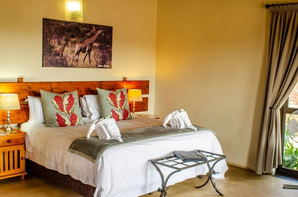 A hotel room with a wooden headboard, a bed with white bedding and decorative pillows, a nightstand with a lamp, and a painting of giraffes on the wall.