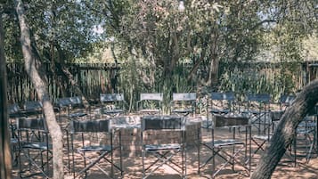 An outdoor seating area with metal chairs and tables arranged in rows, surrounded by trees and a bamboo fence.