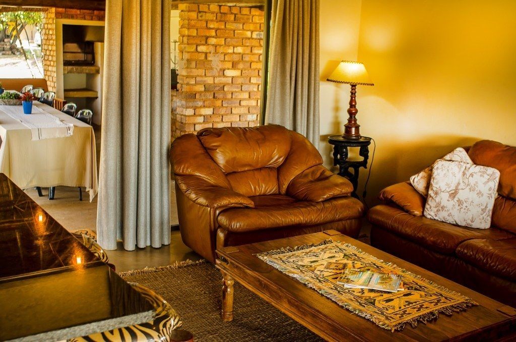 Un salon chaleureux avec un fauteuil en cuir, une table basse en bois et un canapé. On aperçoit un mur en briques et des rideaux au fond.