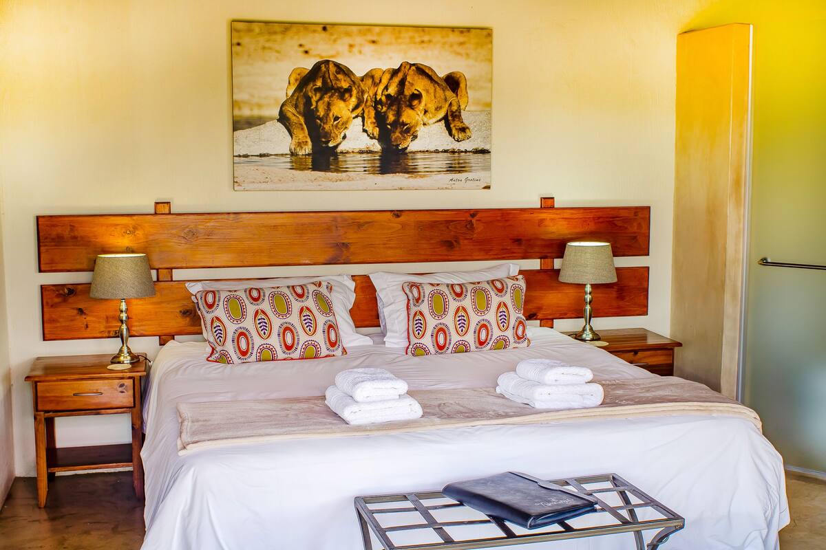 Habitación de hotel con cabecera de madera, almohadas estampadas, cama bien hecha con toallas, mesita de noche con lámpara y un cuadro de elefantes en la pared.