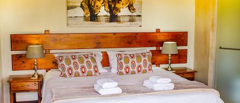 A hotel room with a wooden headboard, patterned pillows, a neatly made bed with towels, a bedside table with a lamp, and a painting of elephants on the wall.