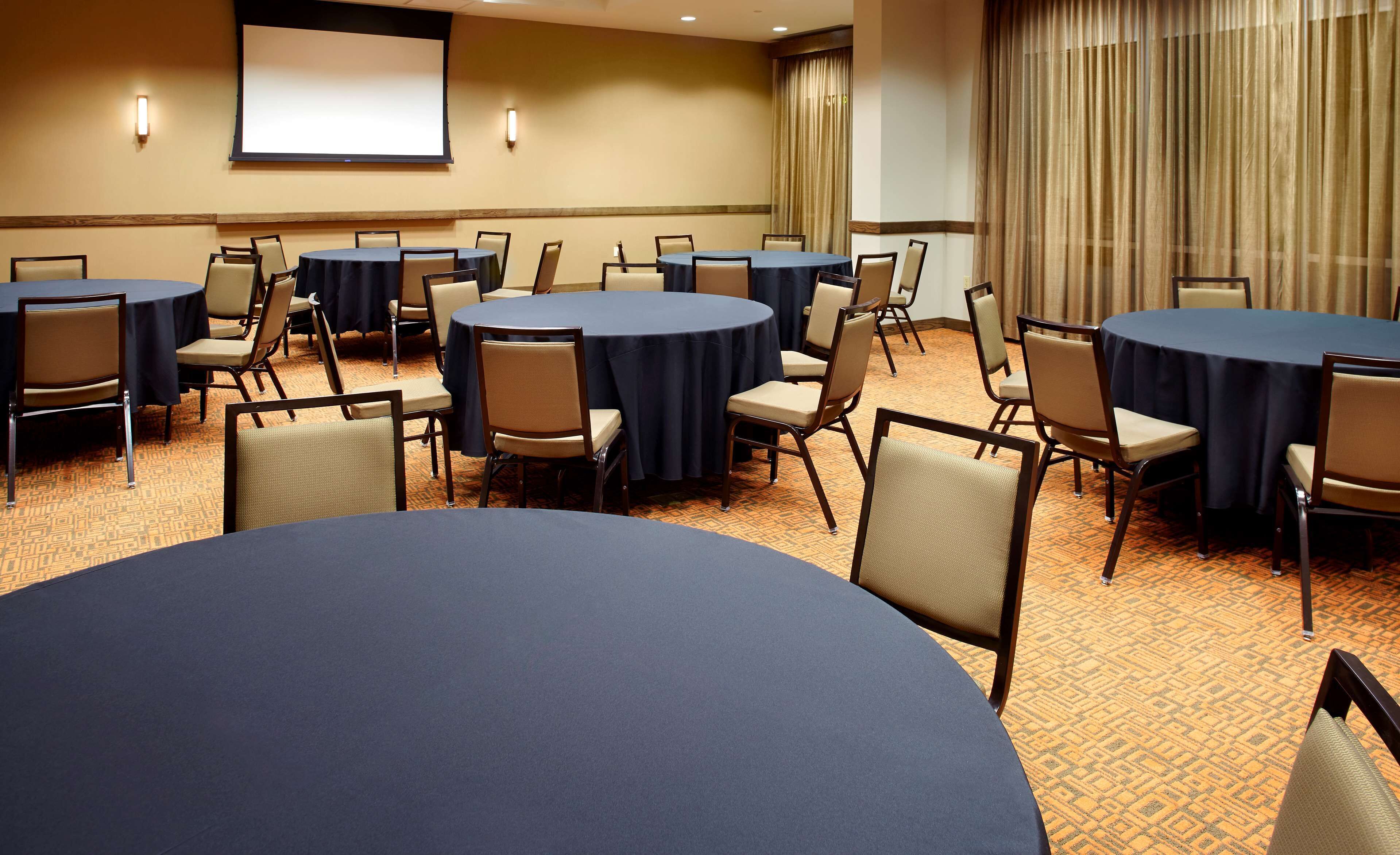 A conference room with multiple round tables, chairs, a projector screen, and a large window with curtains.