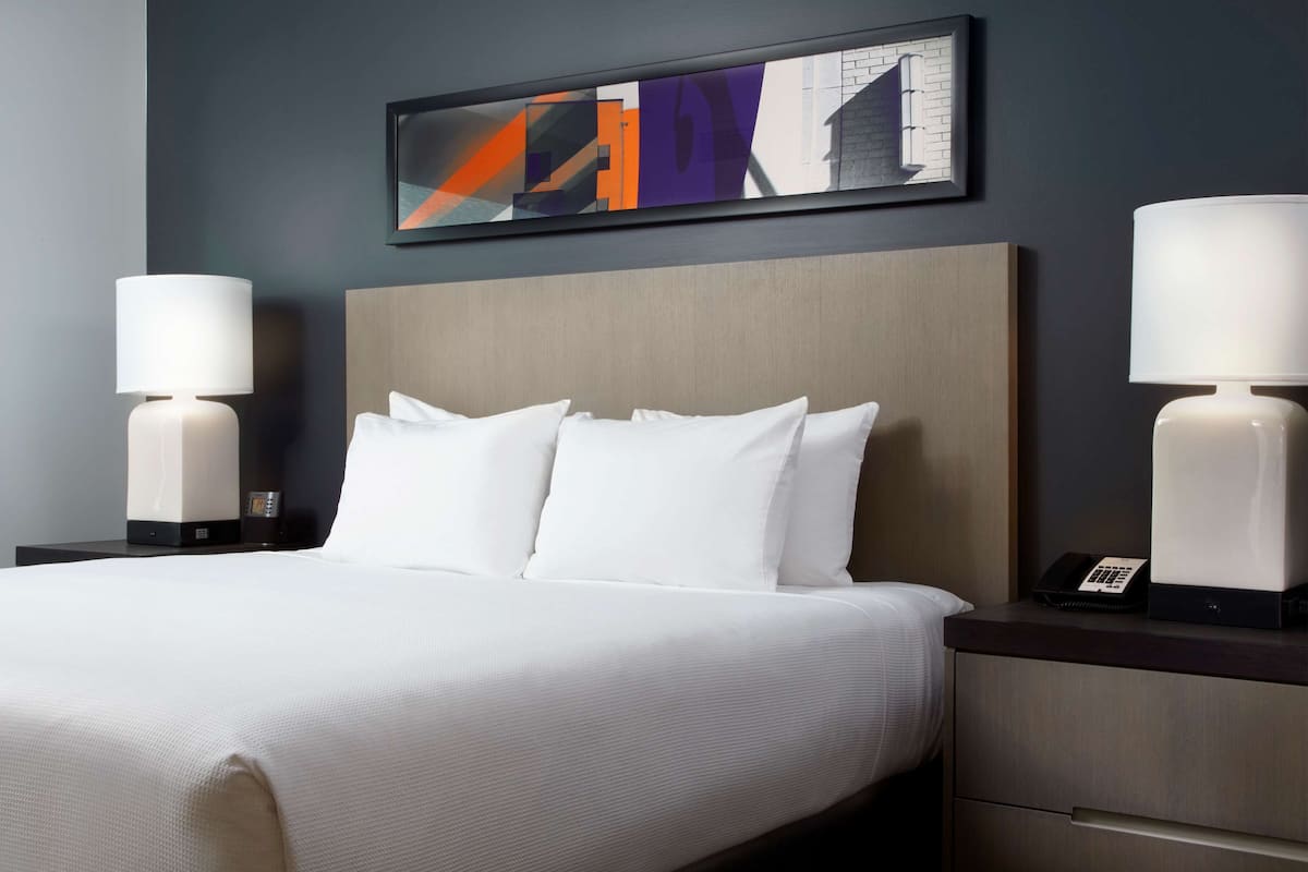 Una habitación de hotel con una cama grande, dos mesitas de noche con lámparas, un cuadro abstracto moderno y un cabecero de madera.