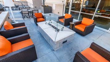 An outdoor patio area with wicker furniture, a glass-top table, and a built-in fire feature.