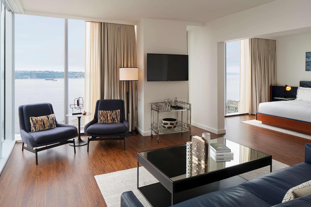 Quarto de hotel moderno com uma grande janela, uma cama, duas poltronas, uma mesa de centro, uma TV e uma mesa lateral com luminária.