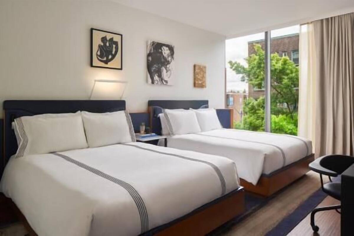 Quarto de hotel com duas camas, uma escrivaninha e vista para o verde lá fora.