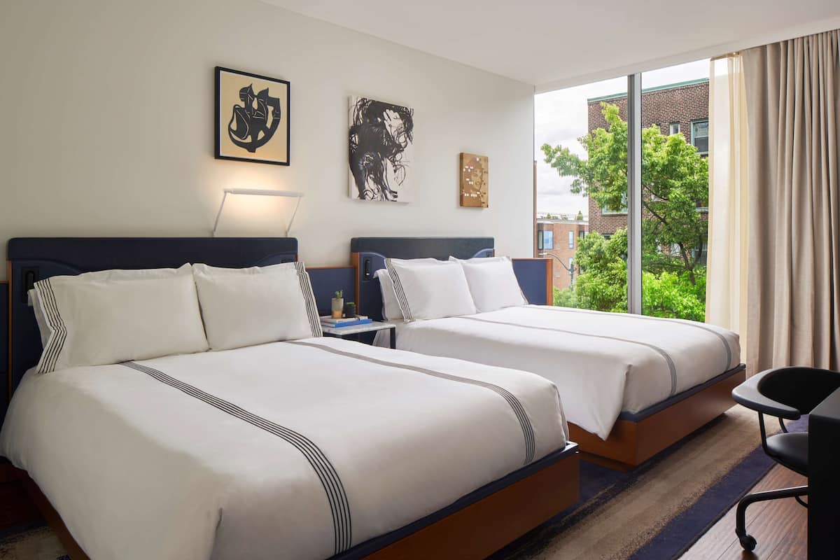 Quarto de hotel com duas camas, uma escrivaninha, uma cadeira e uma janela ampla com vista para edifícios e árvores.