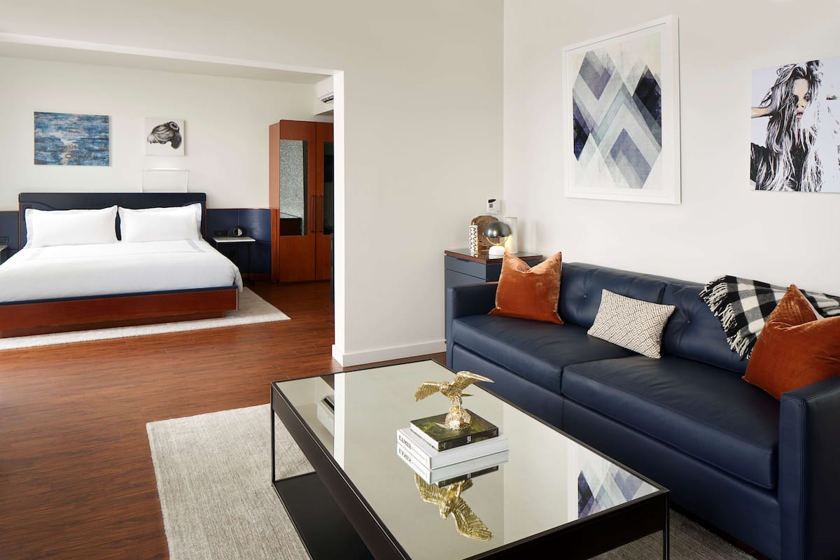 Quarto de hotel moderno com cama, sofá azul e mesa de centro com livros e um objeto decorativo.