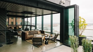 A modern interior with a sofa, chairs, and a dining area, large windows offering a view of a cityscape and a body of water. - Thompson Seattle, by Hyatt (Seattle)