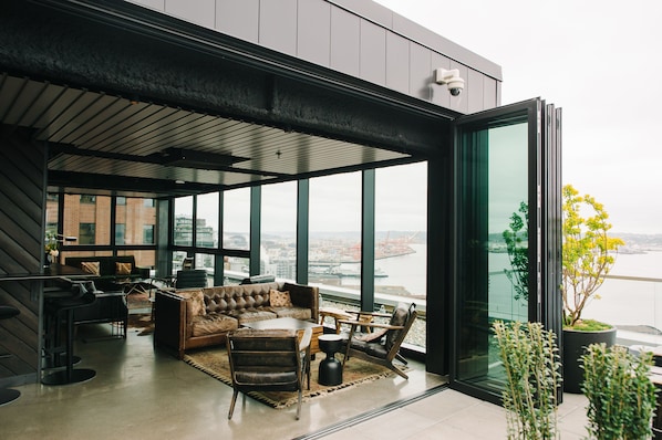 A modern interior with a sofa, chairs, and a dining area, large windows offering a view of a cityscape and a body of water. - Thompson Seattle, by Hyatt (Seattle)