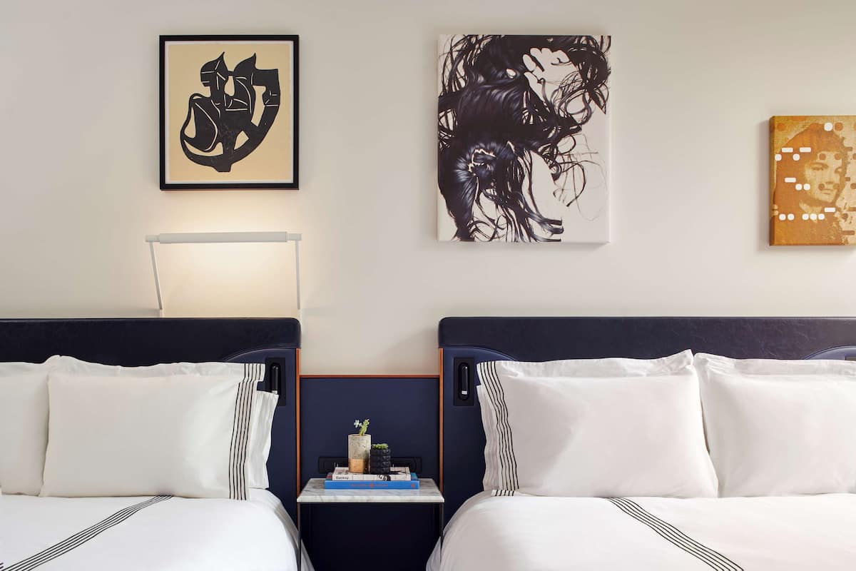 Quarto de hotel com duas camas, uma mesa de cabeceira com abajur e obras de arte nas paredes.