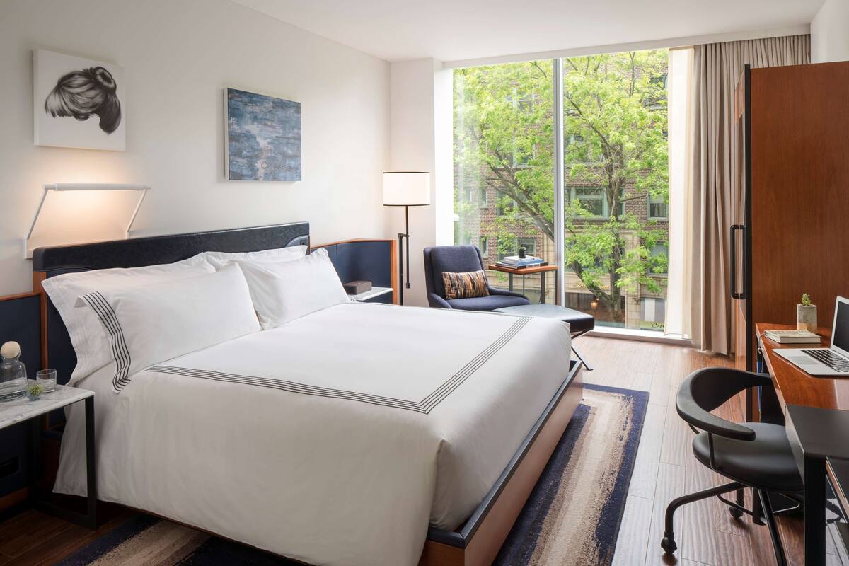 Quarto de hotel com uma cama grande, uma escrivaninha com um laptop, uma cadeira e vista para árvores pela janela.