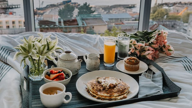 A breakfast tray with pancakes, fruit, and beverages on a bed with a view of a city and water. - Thompson Seattle, by Hyatt (Seattle)