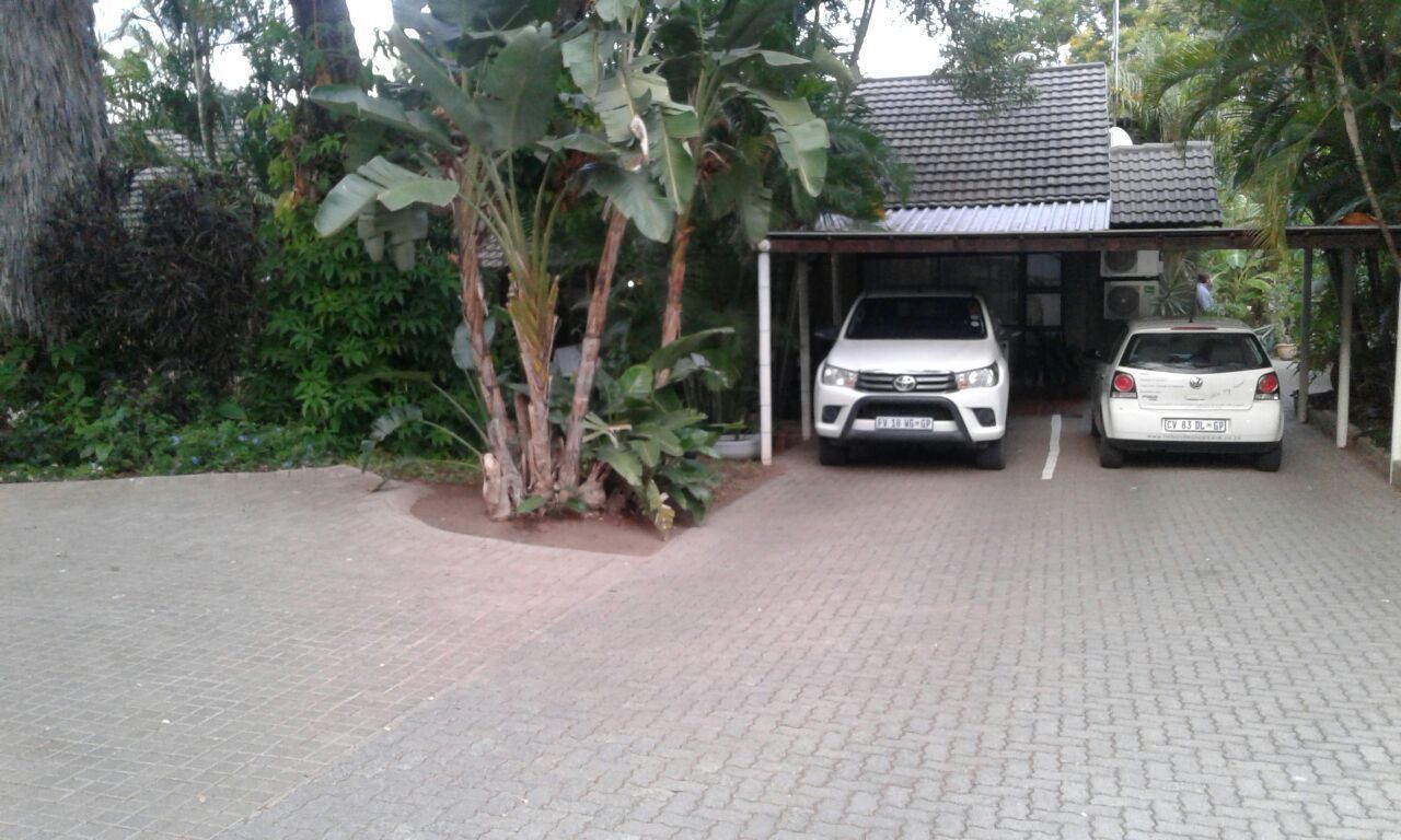 Un área de estacionamiento techada con dos coches aparcados, rodeada de abundante vegetación y una estructura de techo de tejas.
