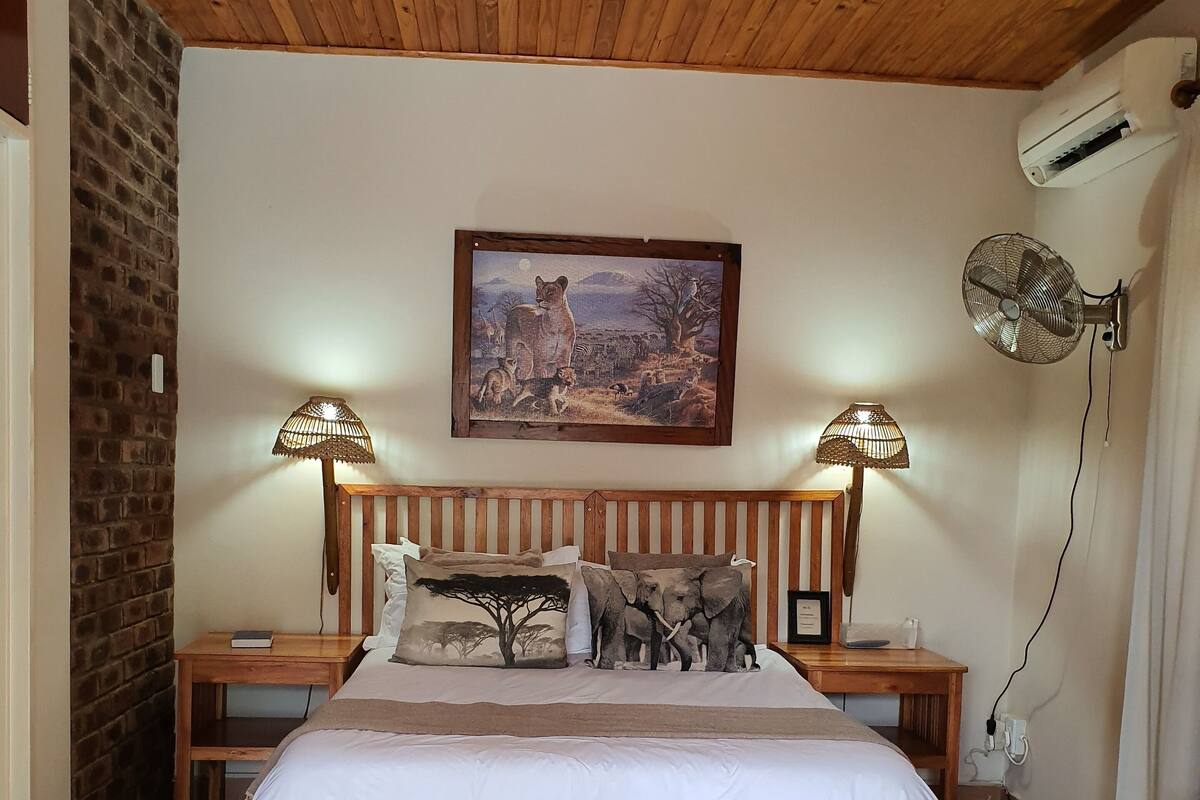 A bedroom with a wooden ceiling, a large bed with white bedding, two bedside tables with lamps, a wall-mounted air conditioner, and a painting of a savannah scene.