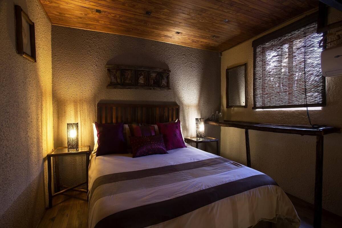A cozy bedroom with a wooden ceiling, a bed with striped bedding, two bedside lamps, and a window with blinds.