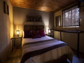 A cozy bedroom with a wooden ceiling, a bed with striped bedding, two bedside lamps, and a window with blinds.