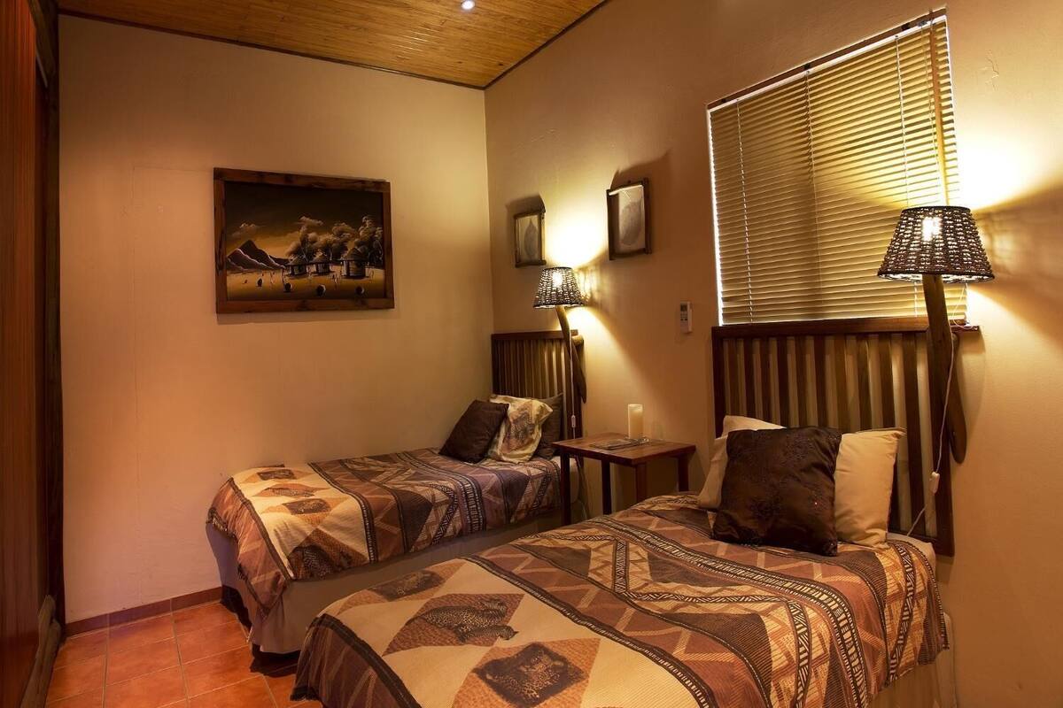 A hotel room with two beds, a painting on the wall, a lamp, and a window with blinds.