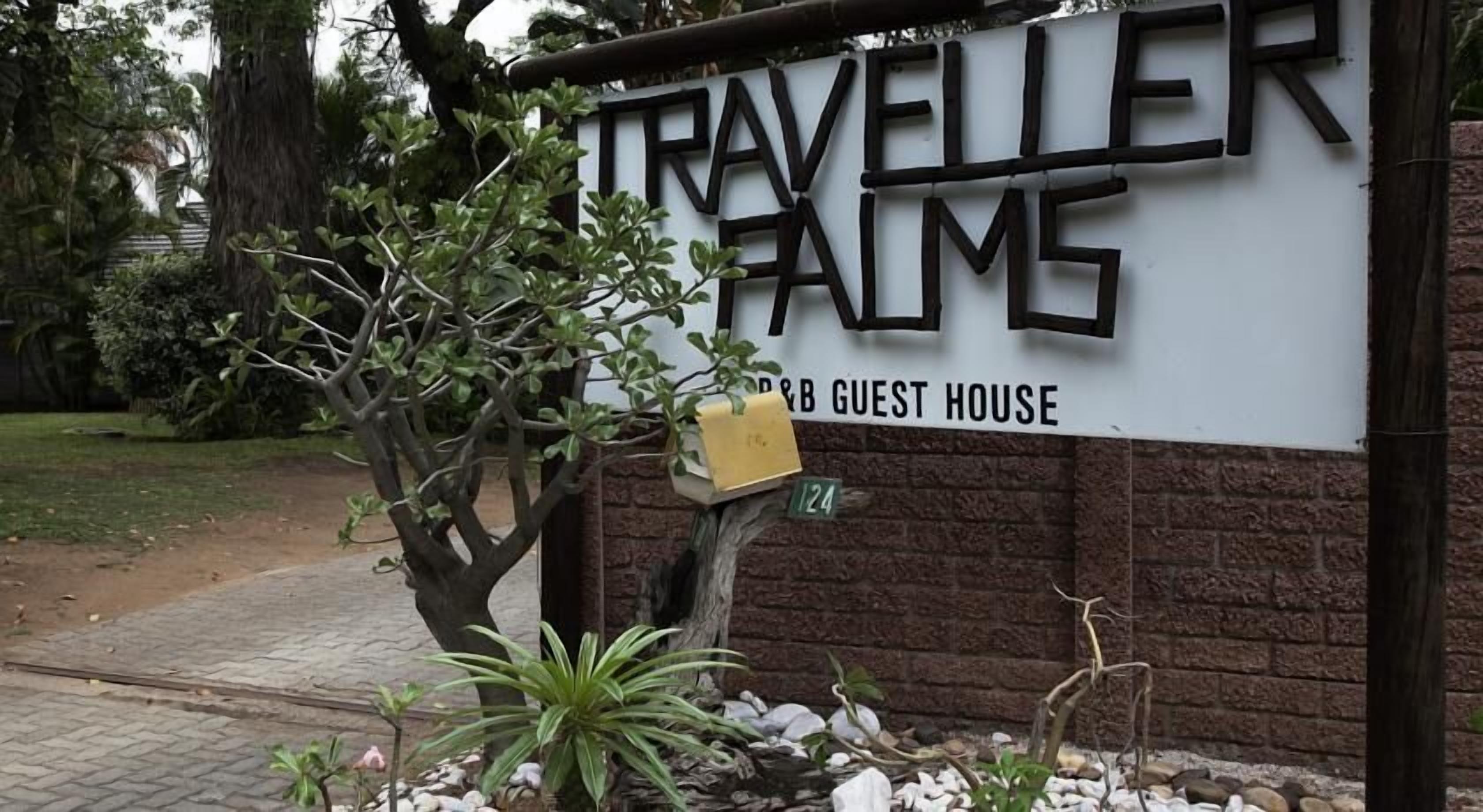 A sign for 'Traveller's Palms' with a guest house label, a tree, and a brick wall.