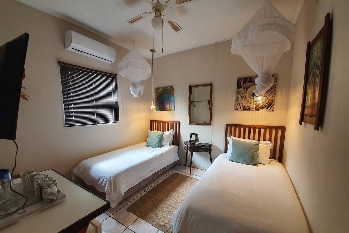 A hotel room with two single beds, a small table with jars, a ceiling fan, an air conditioner, a window with blinds, and a decorative mirror.