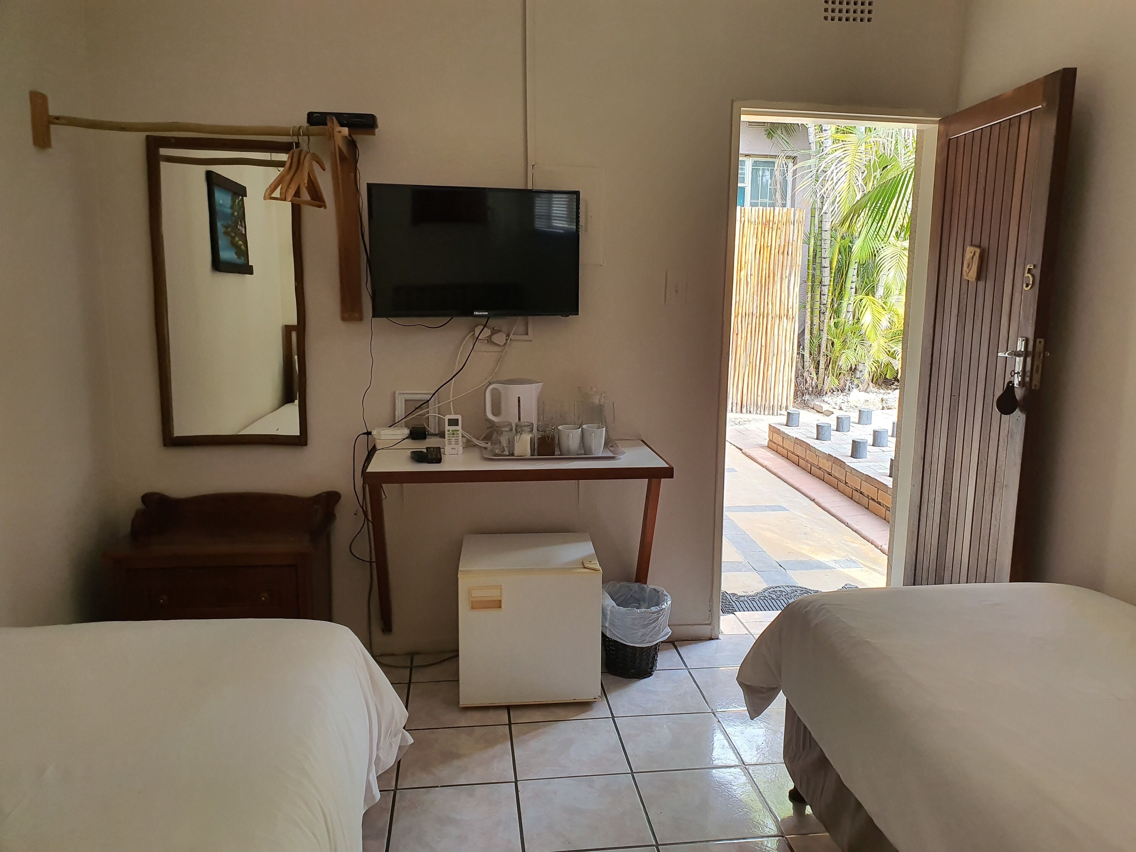 Habitación de hotel con dos camas, un escritorio, televisor, refrigerador pequeño y vista a un jardín desde una puerta abierta.