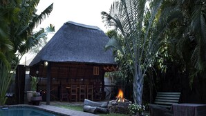 A poolside area with a thatched-roof structure, a fire pit, and lush tropical vegetation. - A Traveller's Palm (Phalaborwa)