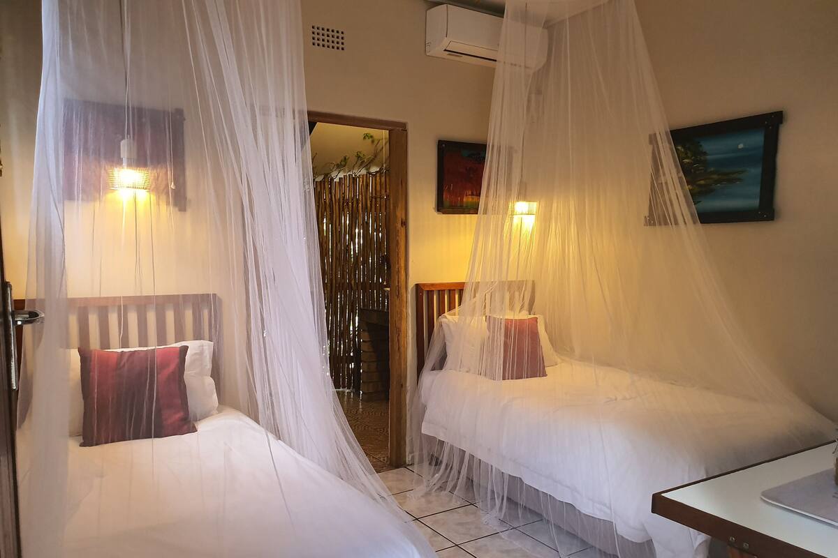 A room with two beds covered by white mosquito nets, a wooden headboard, a small table, and a painting on the wall.