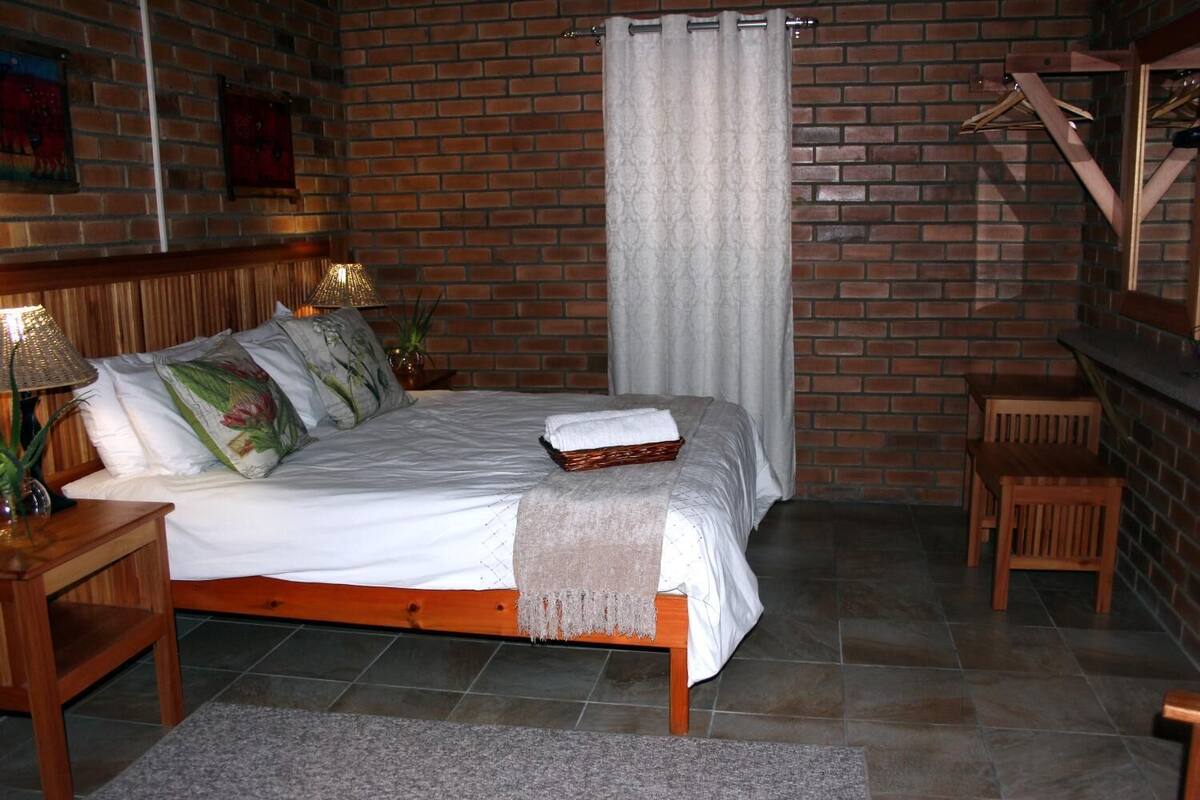 A bedroom with a brick wall, a bed with a white comforter, a wooden headboard, a bedside table with a lamp, and a small wooden chair.