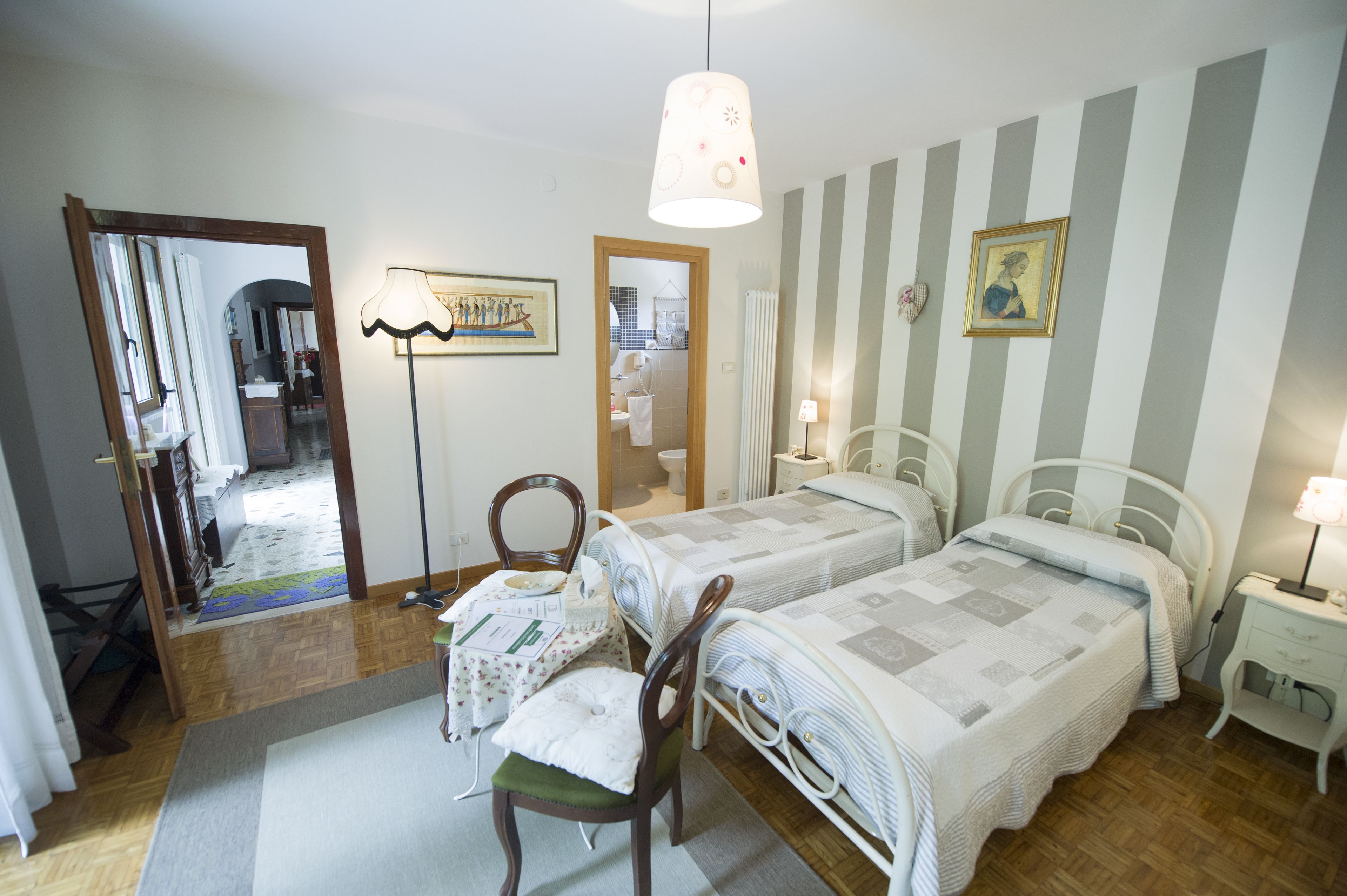 A bedroom with two beds, a chair, a table, a mirror, and a hanging light fixture.