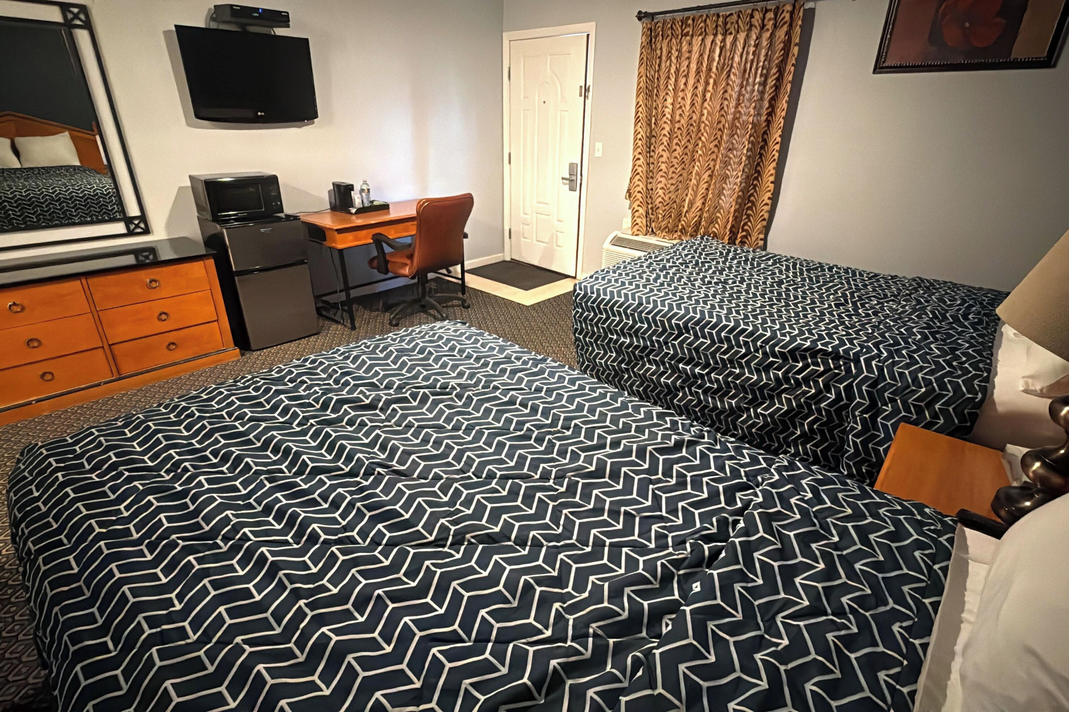 Habitación de hotel con dos camas, un escritorio con silla, televisor, refrigerador y puerta.