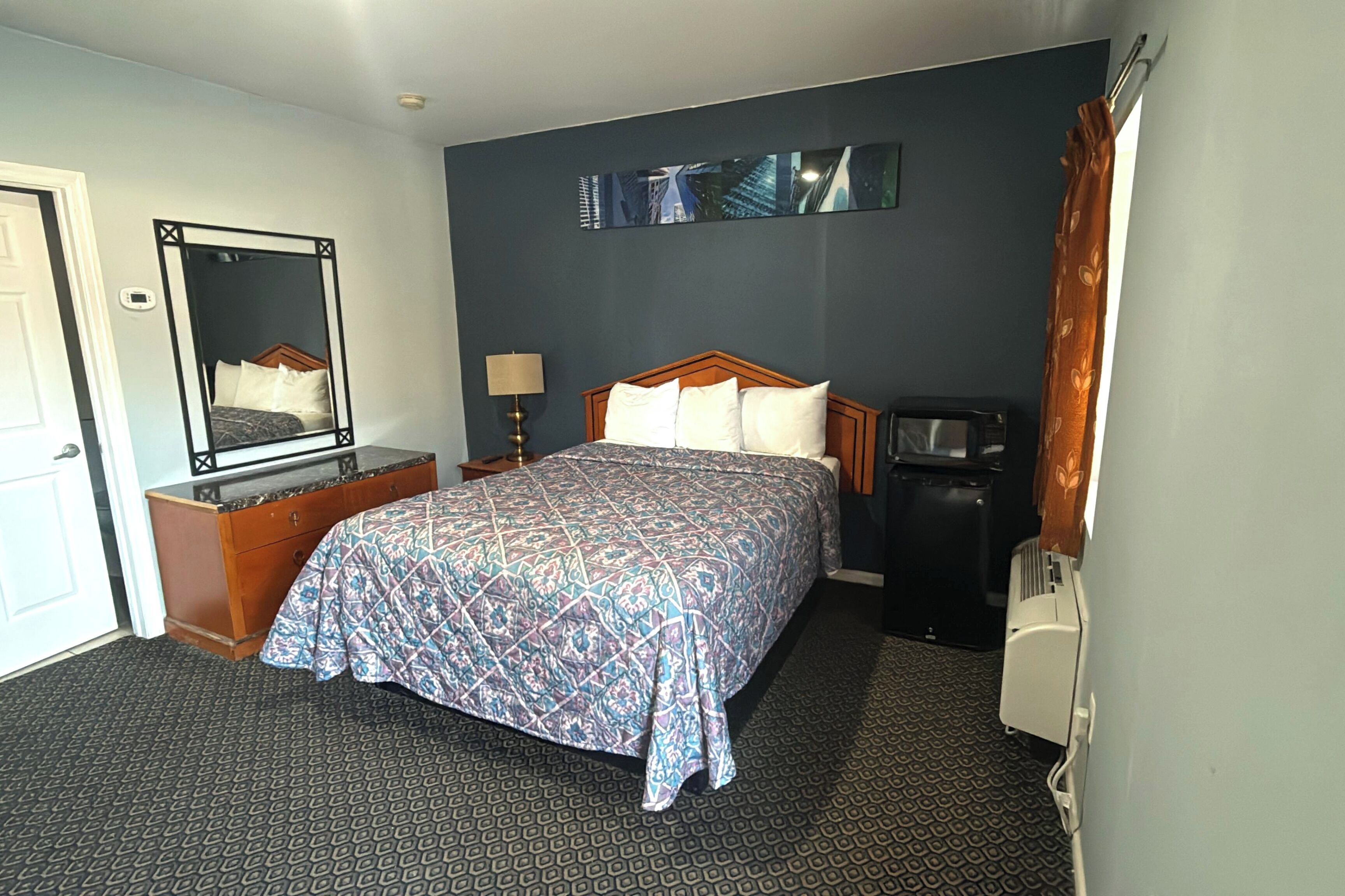 A hotel room with a bed, dresser, mirror, lamp, and a heater.