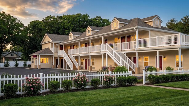 A multi-story hotel with a white picket fence, a covered front porch, and a staircase leading to the upper floors. - At 9 Motel Near Freehold I-195 (Howell)