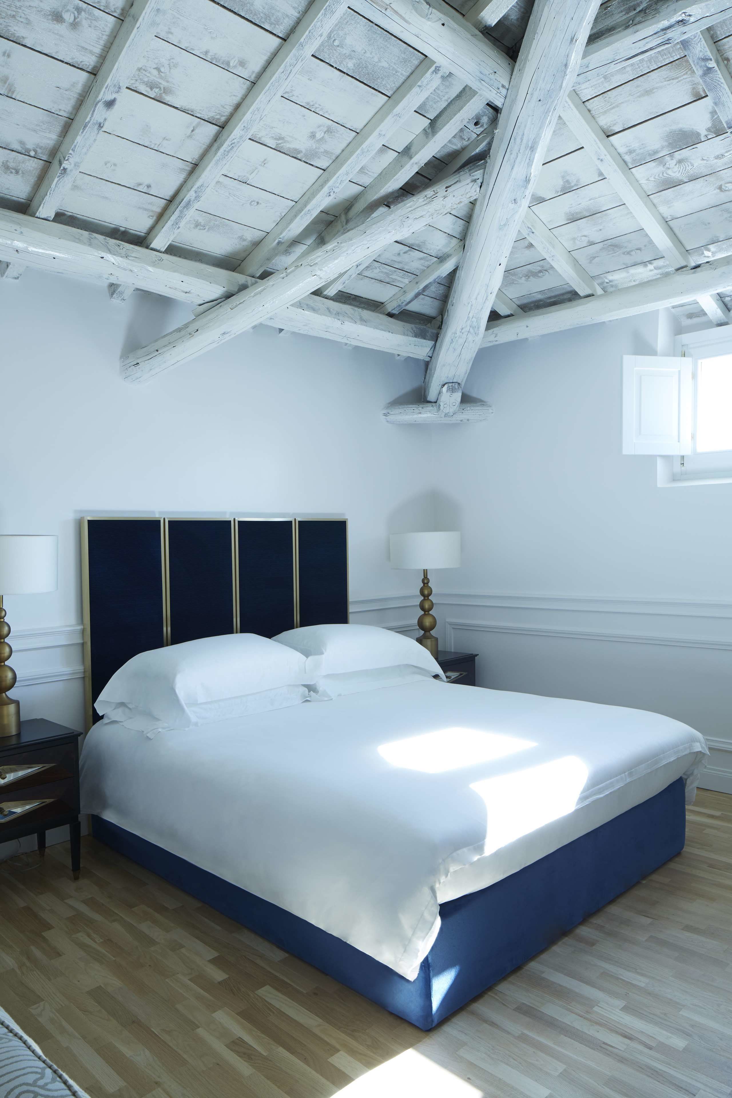 Une chambre avec un grand lit, des poutres en bois au plafond, une table de chevet avec une lampe et une fenêtre avec des rideaux.