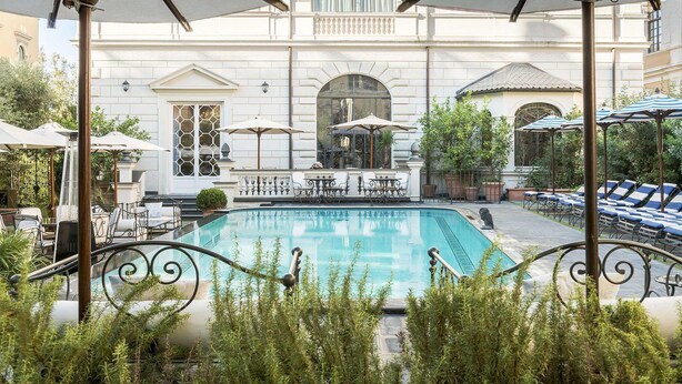 A pool area with lounge chairs and umbrellas, surrounded by a building with arched windows and a fountain. - Palazzo Dama (Rome)