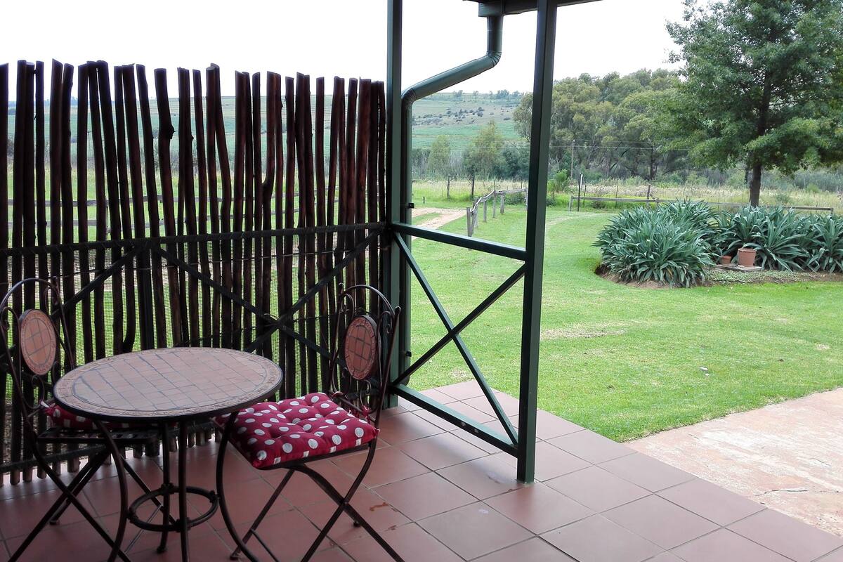 An outdoor patio with a table and chairs, a bamboo fence, and a view of a grassy field with trees.