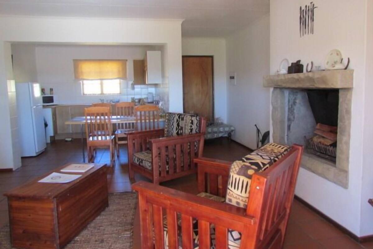 A living room with a fireplace, wooden furniture, a kitchen in the background, and a dining area.