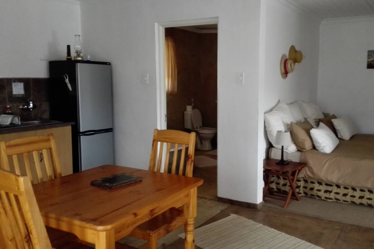 A compact living space with a kitchenette, a dining area with a wooden table and chairs, a bed with patterned bedding, and a small bedside table with a lamp.