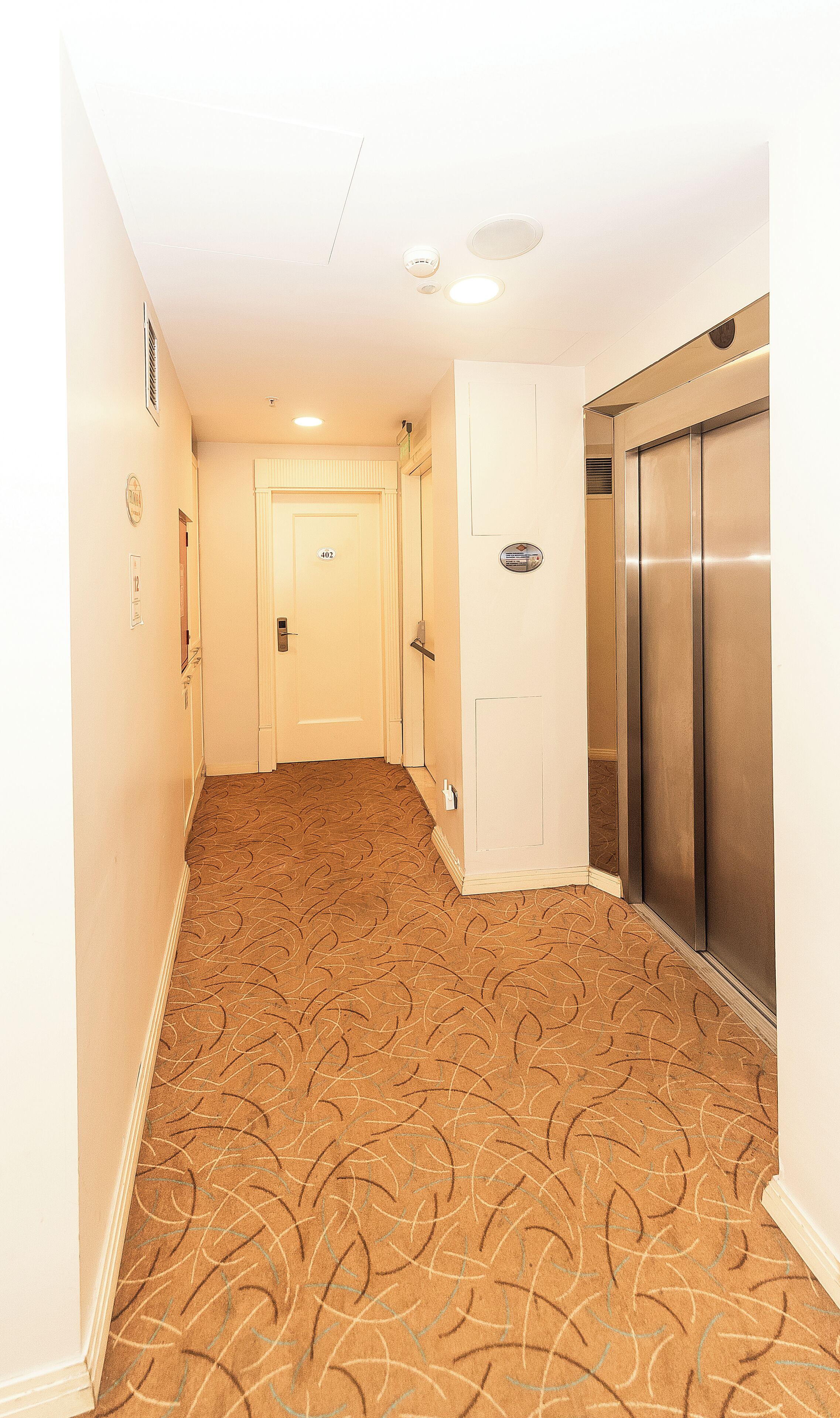 A hotel hallway with patterned carpet, a door marked with the number 402, and a metallic elevator.