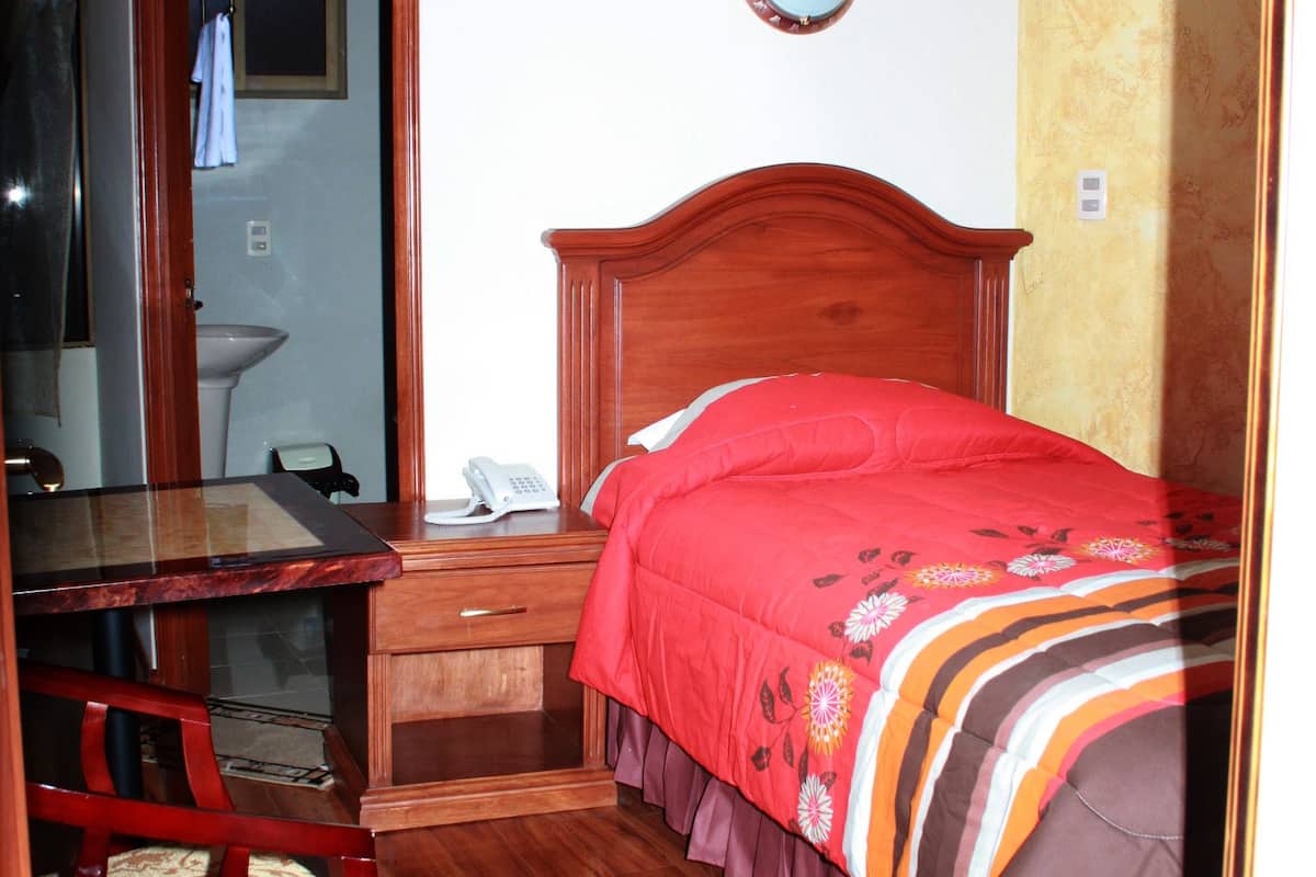 A bedroom with a wooden bed, a bedside table, a chair, a mirror, and a bathroom visible through an open door.