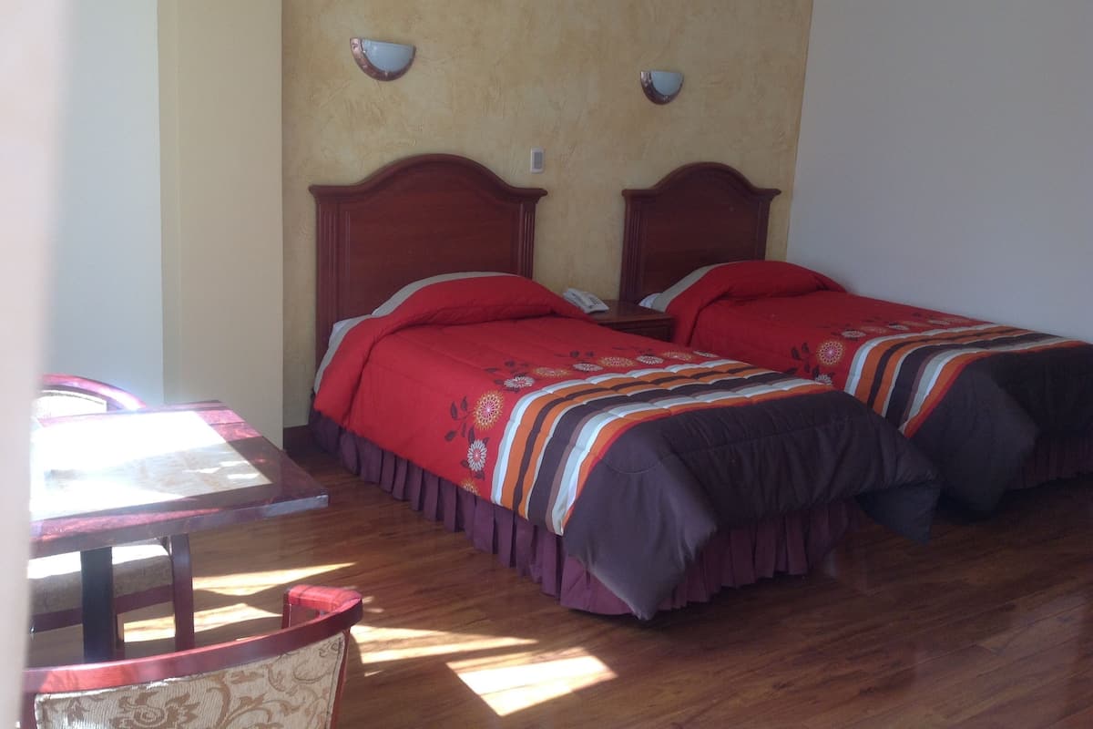 A room with two wooden beds, each with a red bedspread and a wooden headboard. There is a small table with a chair in the foreground, and a wall-mounted light fixture above the beds.