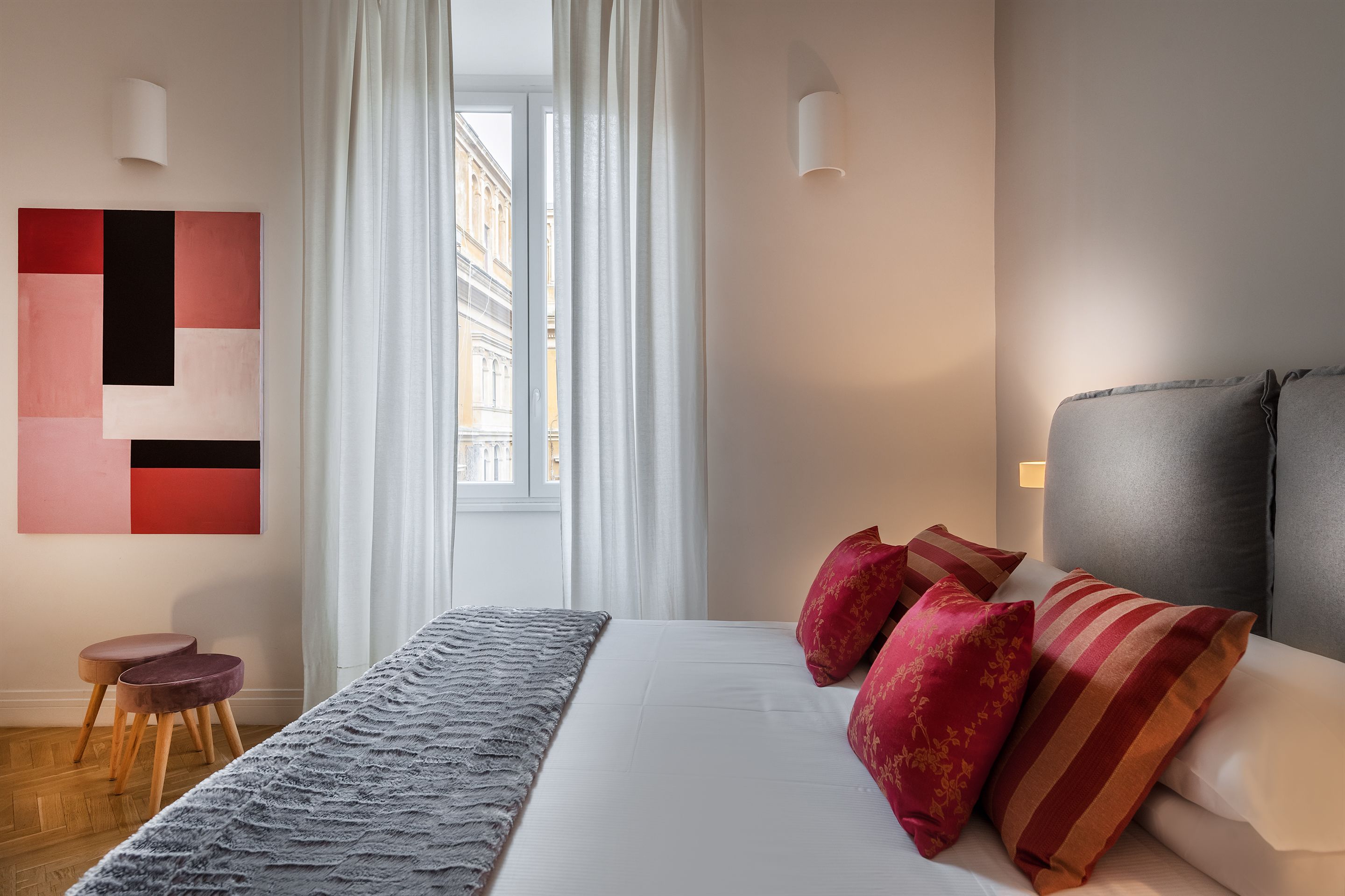 A modern hotel room with a large bed, red and orange pillows, a grey throw, and a view of buildings through sheer curtains.