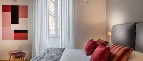 Una habitación de hotel moderna con una cama grande, almohadas rojas y anaranjadas, una manta gris y vistas de edificios a través de cortinas translúcidas.