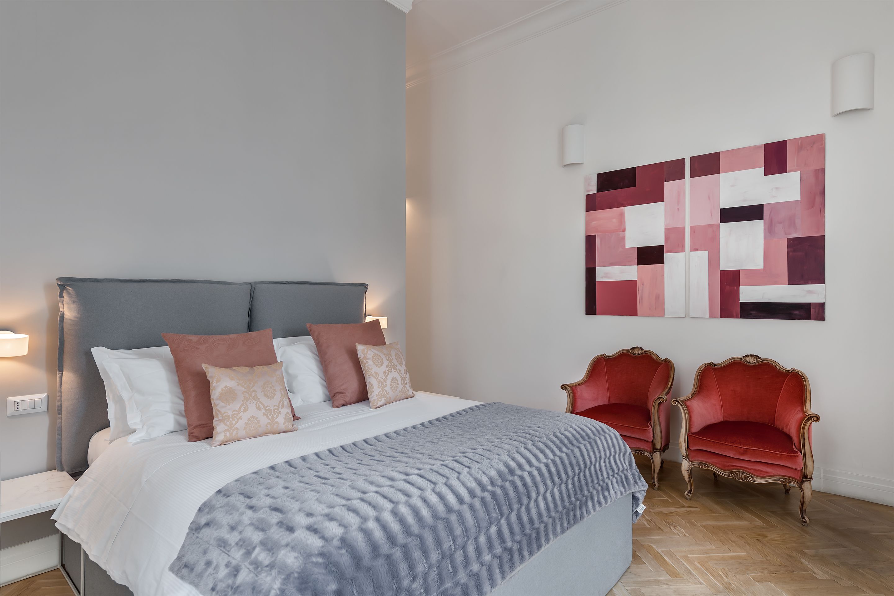 A neatly made bed with a grey and white duvet, two red upholstered chairs, and a geometric abstract painting on the wall.