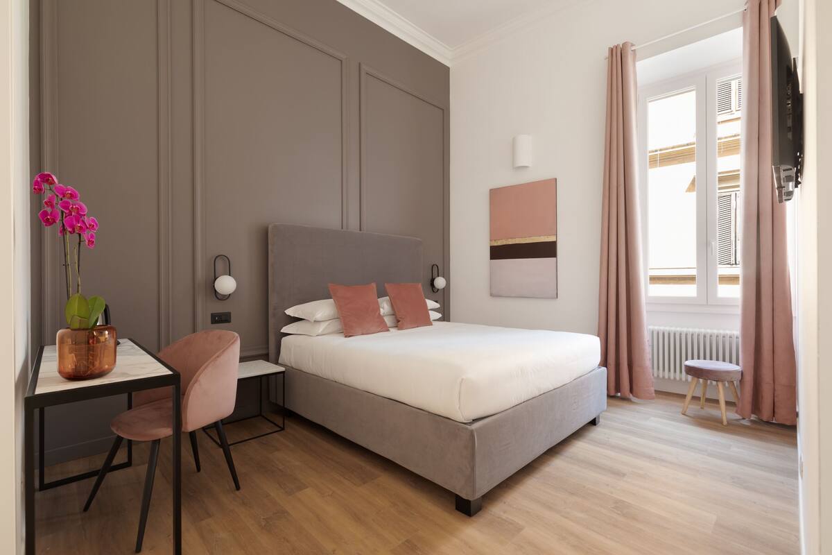 Una habitación de hotel moderna con una cama, un escritorio con una silla, un taburete pequeño, una planta en maceta y una ventana con cortinas.